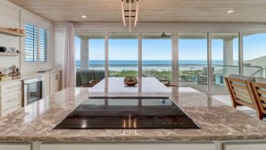 Cozinha moderna com uma ilha grande, armários brancos e vista para a praia.