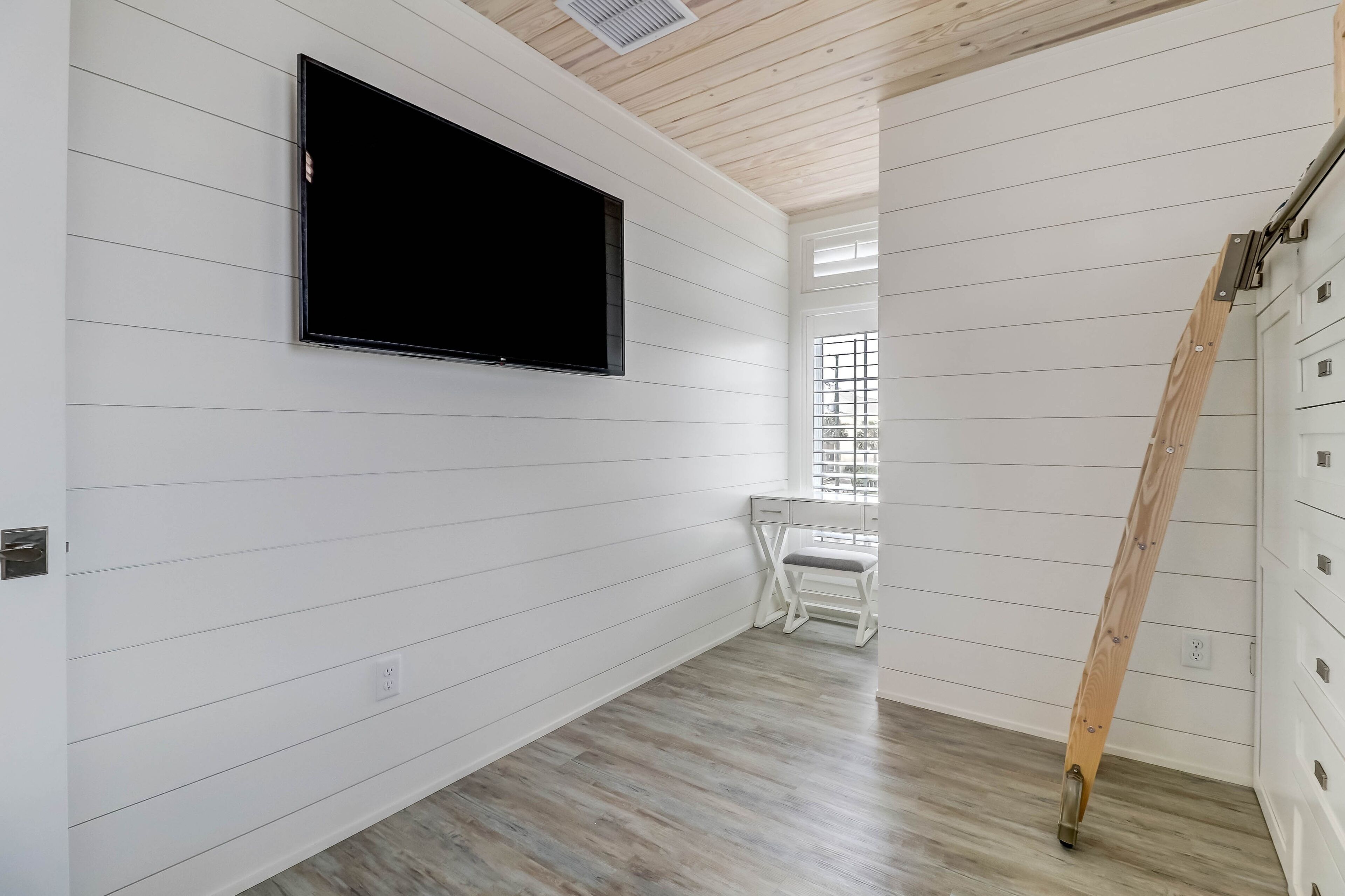 A room with white paneled walls, a mounted flat-screen TV, a wooden chair, and a wooden ladder leaning against the wall.