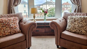 Une pièce comprenant deux fauteuils en cuir, une petite table avec une lampe et un vase, et une fenêtre donnant sur des immeubles.