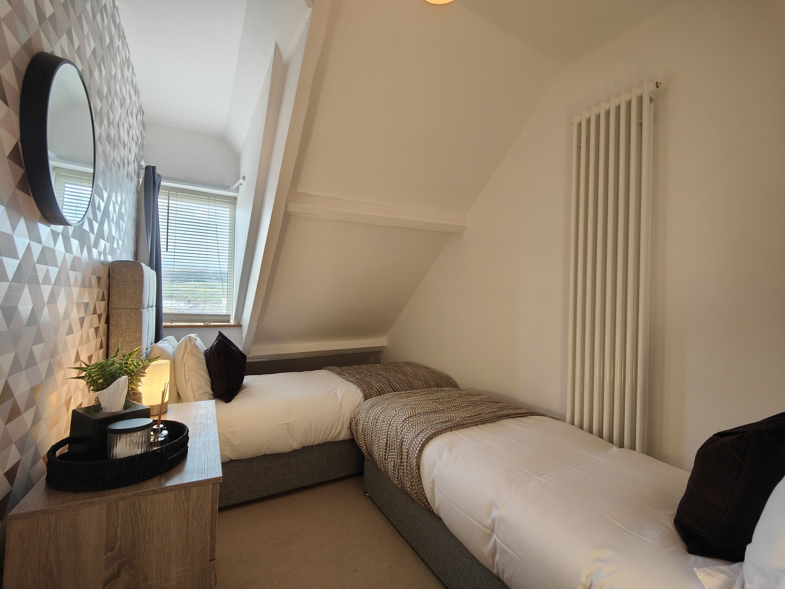 A bedroom with two beds, a bedside table with a lamp, a mirror, and a radiator.