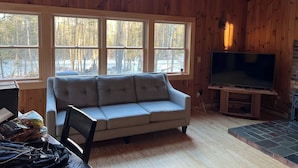 Un intérieur de chalet chaleureux avec un canapé, une télévision et de grandes fenêtres donnant sur une forêt enneigée.