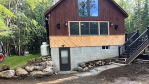 Une cabane en bois à deux étages, avec des fondations en béton, une terrasse en bois et un escalier menant à l’étage supérieur.
