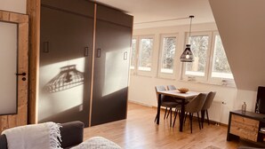 Ein modernes Wohnzimmer mit einer Couch, einem Esstisch mit Stühlen, einem großen Fenster mit Blick auf Bäume und einer Deckenleuchte.