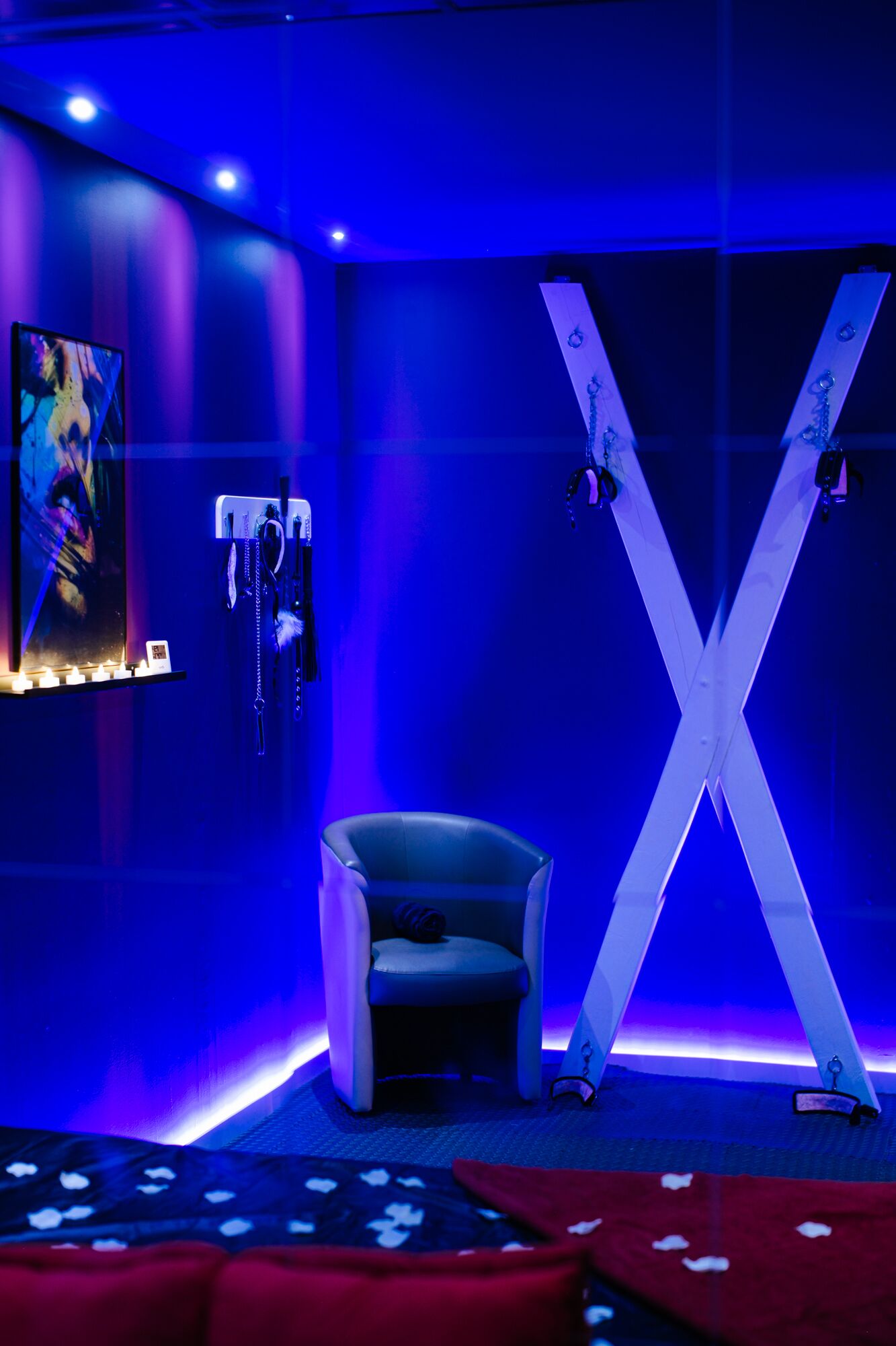 A room with a blue-lit ambiance, featuring a large X-shaped structure, a chair, and wall-mounted jewelry.