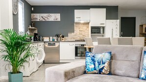 Ein modernes Wohnzimmer mit einem grauen Sofa, einem blauen Couchtisch und einem weißen Beistelltisch. In der Ecke steht eine große Topfpflanze.