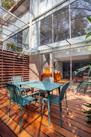 En moderne udendørs terrasse med et glasindrammet spiseområde, træterrasse og udsigt til de omkringliggende træer.