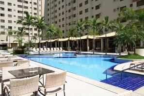 A swimming pool with lounge chairs and tables, surrounded by palm trees and a multi-story building.