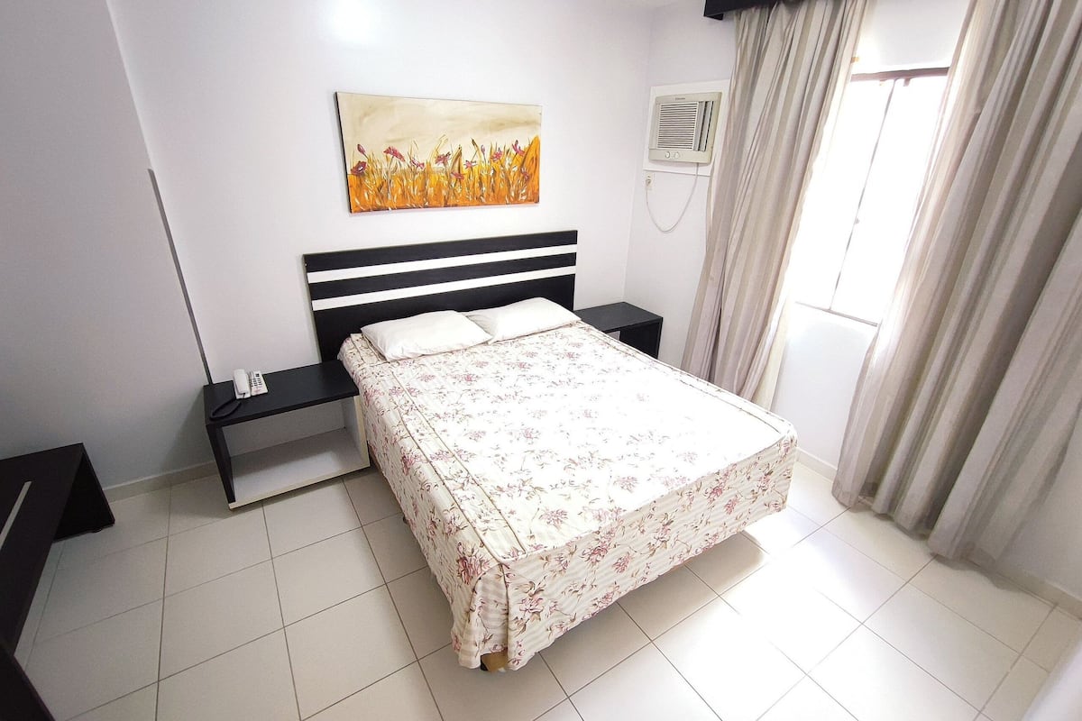 A bedroom with a bed, a bedside table, a phone, a painting, an air conditioning unit, and a window with curtains.