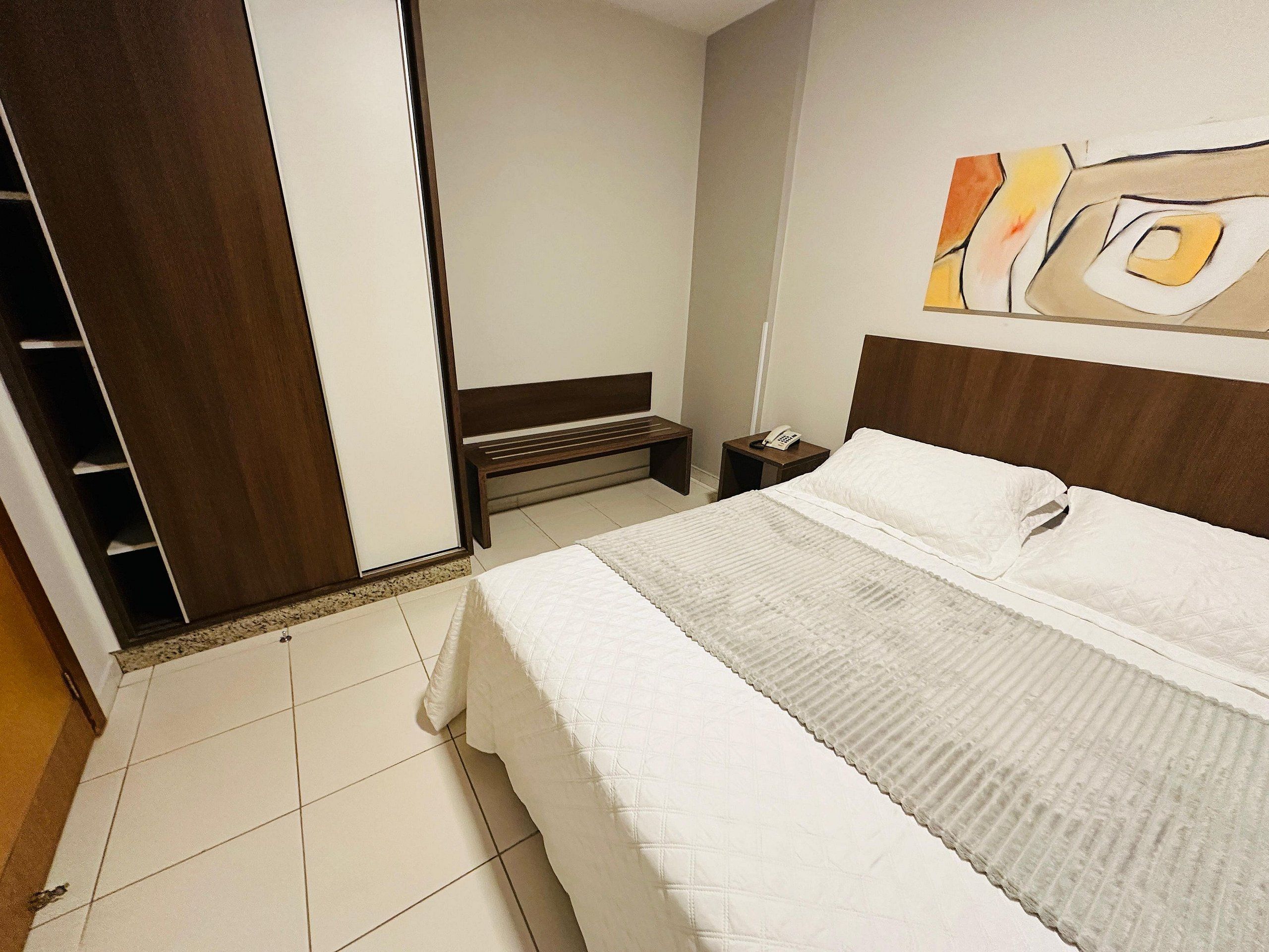 Een hotelkamer met een bed, nachtkastje met telefoon, een bankje en een kledingkast.