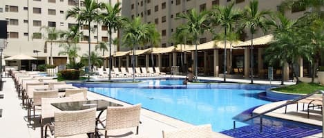A swimming pool with lounge chairs and umbrellas, surrounded by palm trees and a multi-story building.