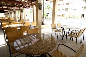 A covered outdoor seating area with round tables and wicker chairs, a pool in the background, and a building with multiple windows. - Thermas Boulevard (Caldas Novas)