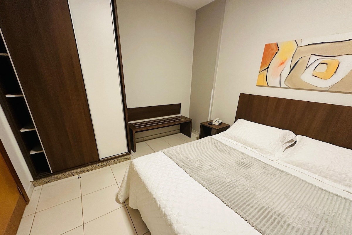 A hotel room with a bed, a bedside table with a phone, a bench, and a wardrobe.