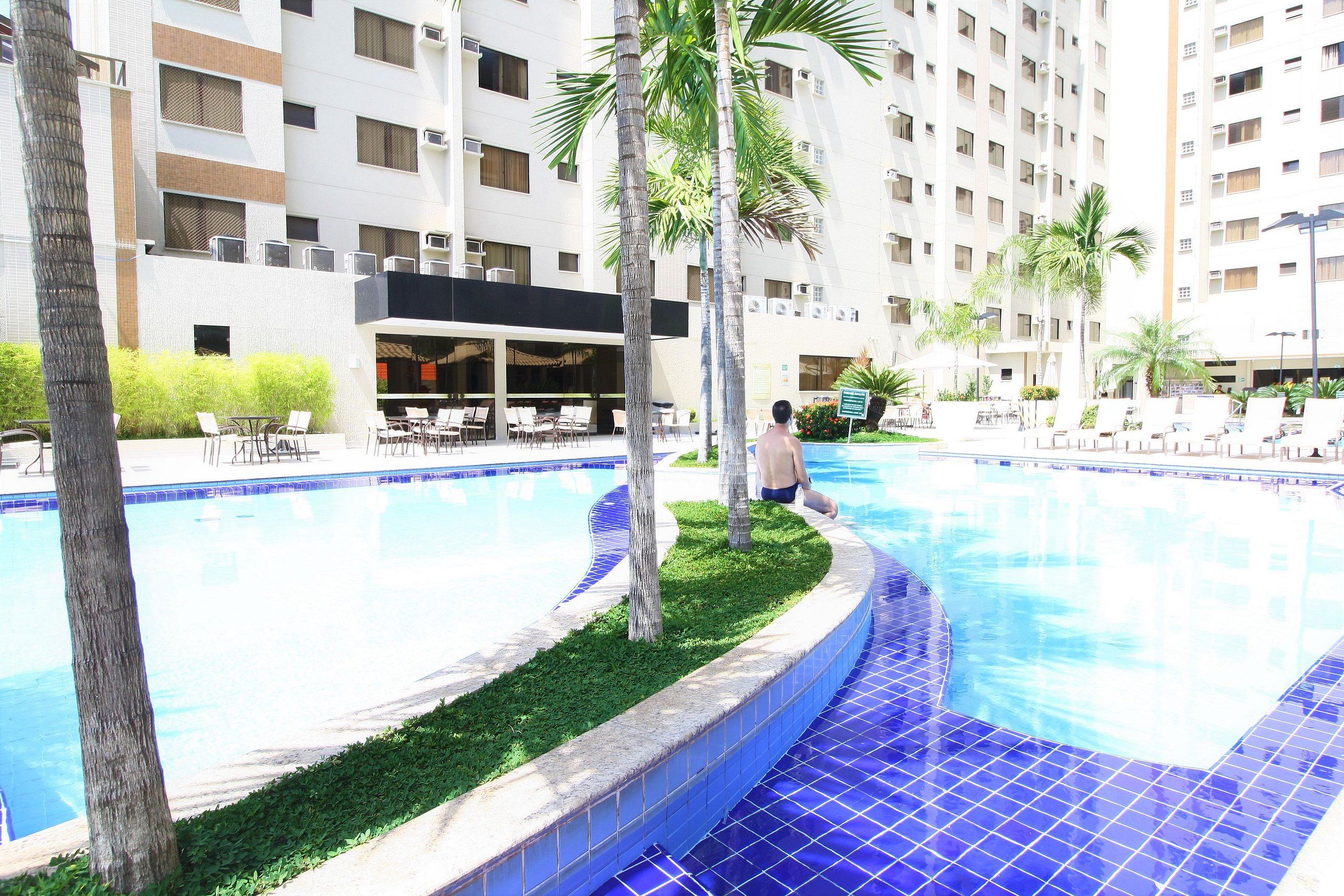 Una piscina con sillones de descanso y palmeras, rodeada por un edificio de hotel de varios pisos.