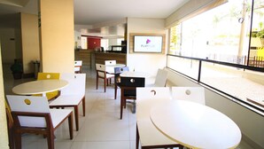 A dining area with white tables and chairs, a PlayPlus screen, and a view of the street through large windows. - Thermas Boulevard (Caldas Novas)