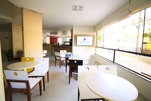 A dining area with white tables and chairs, a PlayPlus screen, and a view of the street through large windows. - Thermas Boulevard (Caldas Novas)