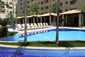 A swimming pool with lounge chairs, palm trees, and a building with multiple floors in the background.
