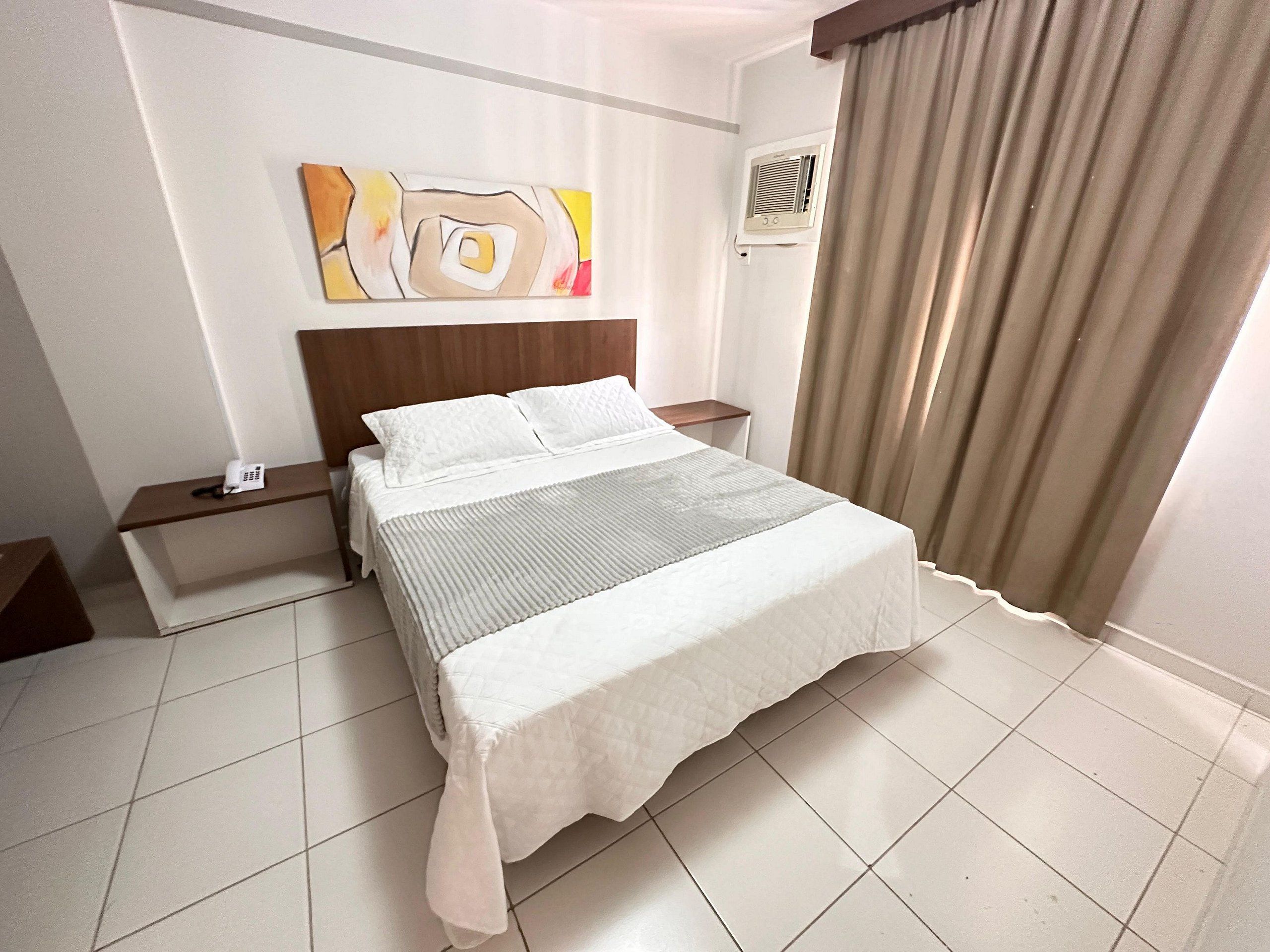 Una camera d'albergo con un letto, un comodino, un telefono, un quadro appeso al muro e una finestra con tende.