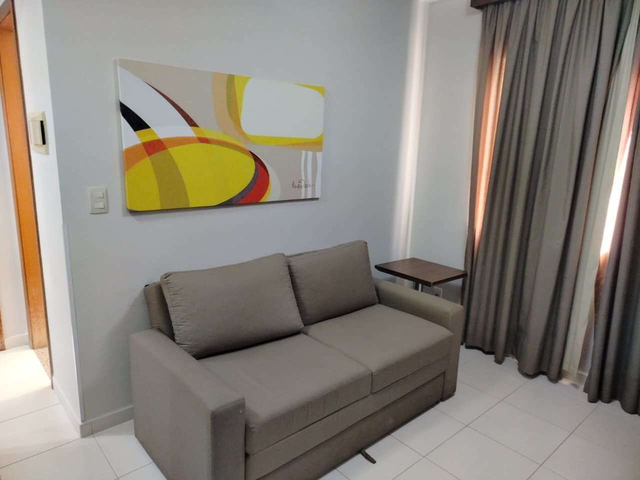 Una sala de estar con un sofá, una mesita, un cuadro en la pared y una ventana con cortinas.