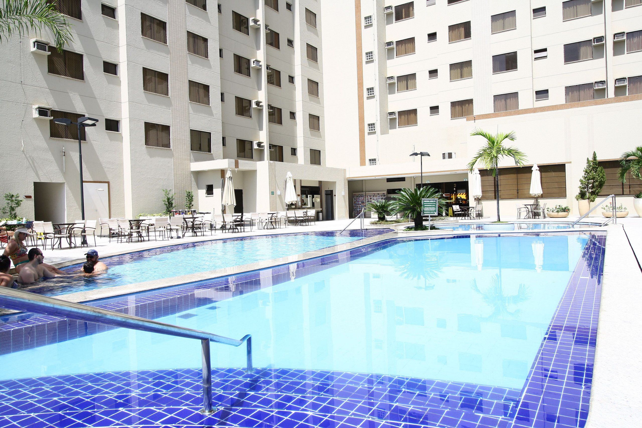 A swimming pool with tiled blue floors, surrounded by lounge chairs and umbrellas, with a multi-story building in the background.