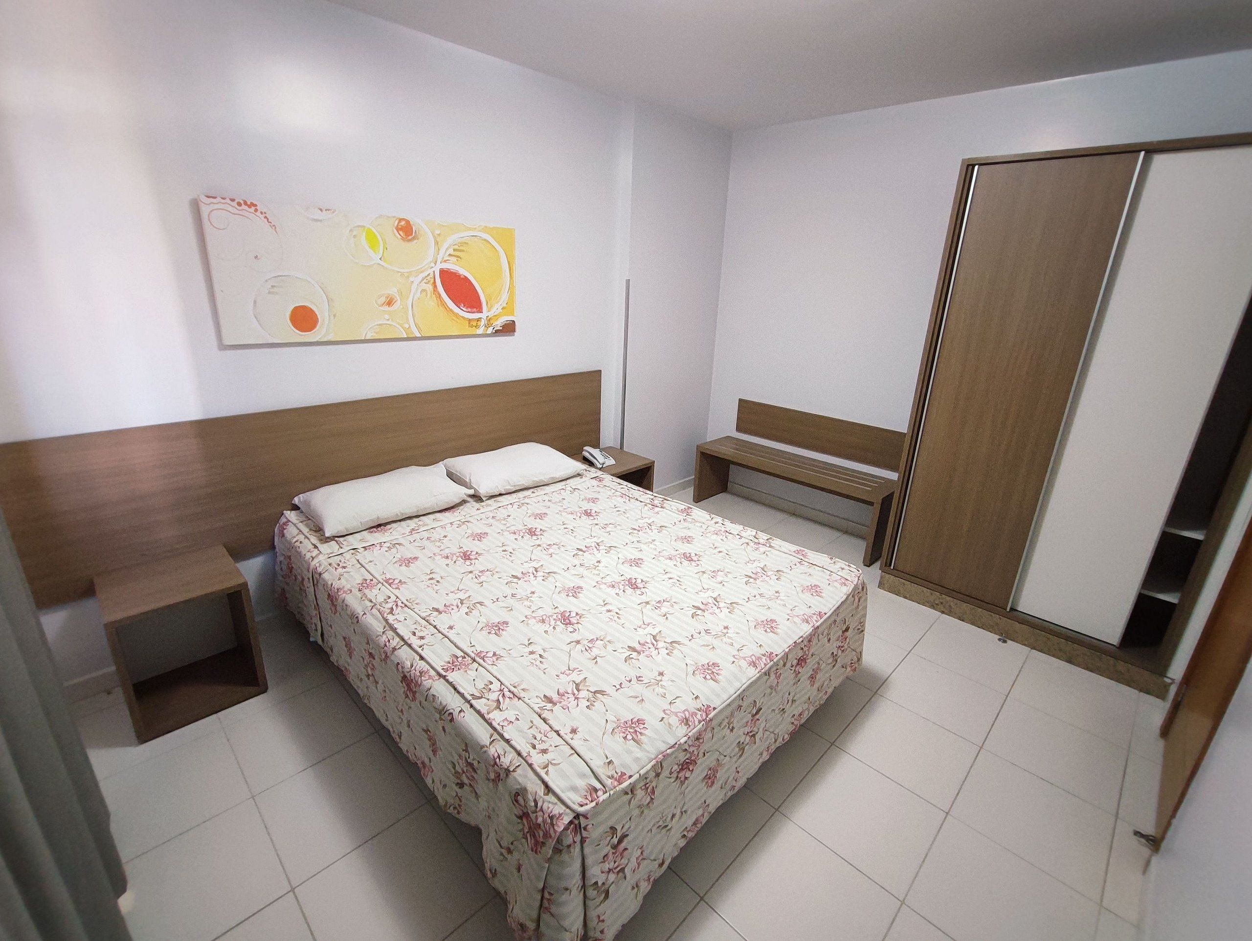 Una habitación de hotel con cama, mesitas de noche, un banco, armario y una obra de arte en la pared.