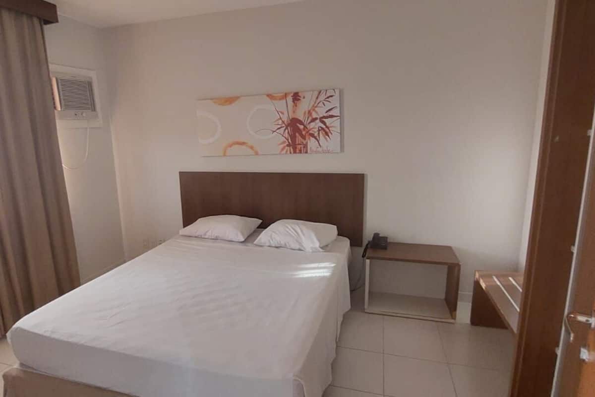 A hotel room with a bed, a bedside table, a wall-mounted phone, and a window with curtains.
