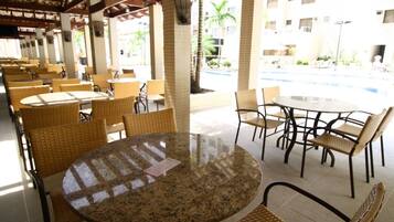 A covered outdoor seating area with round tables and wicker chairs, a pool in the background, and a building with multiple windows.