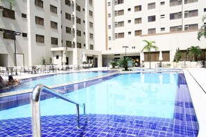 A swimming pool with blue tiles, surrounded by outdoor seating and palm trees, with a multi-story building in the background.