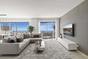 A modern living room with a large sectional sofa, a glass coffee table, and a flat-screen TV mounted on the wall.