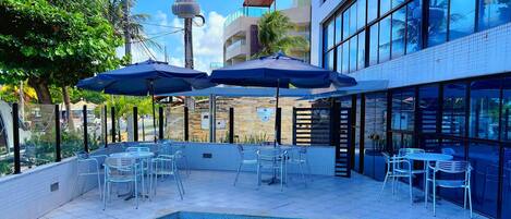 Zona de piscina al aire libre con sillas de descanso y sombrillas, rodeada de edificios con grandes ventanales y un cielo despejado.