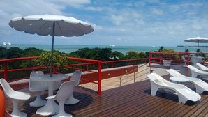 Eine Terrasse mit weißen Plastikstühlen und -tischen, roten Geländern und Blick auf den Ozean.