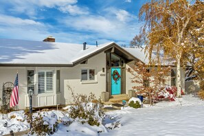 Una casa de un piso con puerta azul, una chimenea y un jardín cubierto de nieve.