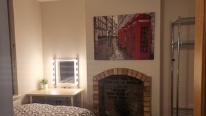 A bedroom with a brick fireplace, a desk with a mirror, a red telephone booth painting, and a metal shelving unit.