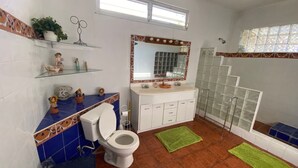 Baño con inodoro, lavabo y ducha. Hay objetos decorativos en los estantes y una ventana sobre el lavabo.
