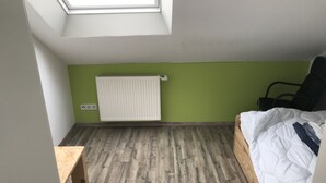 A small, well-lit room with a sloped ceiling, a radiator, a bed, and a wooden table. - Single room (Homberg (Efze))