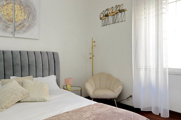 A bedroom with a bed, pillows, a bedside lamp, a chair, a wall-mounted light fixture, and a window with curtains. - Domus Lou Luxury Apartment in Trastevere Rome (Roma)