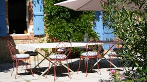 Un área de comedor al aire libre con una mesa, sillas, un paraguas blanco y vistas a un edificio con contraventanas azules.