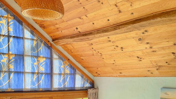 Un dormitorio con techo de madera, un ventanal con vistas a la vegetación, una cama con ropa de cama estampada y una mesita de noche con una lámpara.