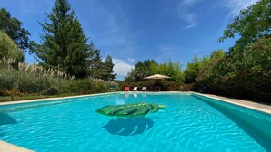 Una piscina azul cristalina con un juguete inflable verde, rodeada de árboles y una zona de estar cubierta.