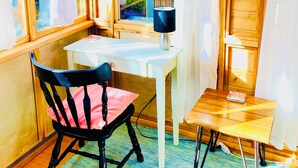 Una habitación acogedora con paredes de madera y un ventanal con vistas a un jardín. Hay una mesa con una silla, una mesita de noche con una lámpara y una ventana con cortinas.