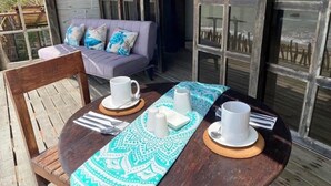 Une table en bois, dressée avec des tasses blanches et une nappe à motifs, un canapé confortable agrémenté de coussins décoratifs, et une vue sur l’océan par de grandes fenêtres.