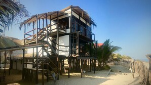 Un immeuble à plusieurs étages, doté d’un escalier en bois et d’un toit de chaume, entouré de palmiers et bordé d’une plage de sable.