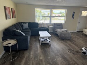 Una sala de estar con un sofá seccional, una mesita, un sillón y una ventana con persianas.
