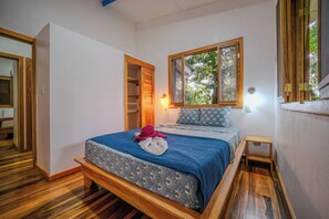 A bedroom with a bed, a nightstand, a window with a view of trees, and a door leading to another room.