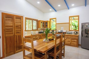 A kitchen with wooden cabinets, a dining table, chairs, a refrigerator, and a window with a view of greenery.
