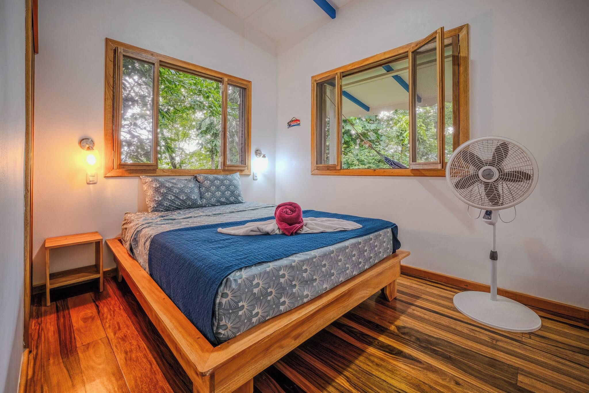 Un dormitorio con una cama de madera, un ventilador y dos ventanas con vistas a la vegetación.