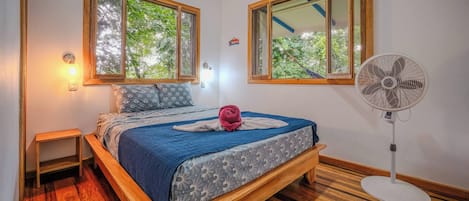 A bedroom with a wooden bed, a standing fan, and two windows with a view of greenery.