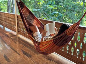 Iemand ontspant in een hangmat op een houten terras met een leuning, en leest een boek.
