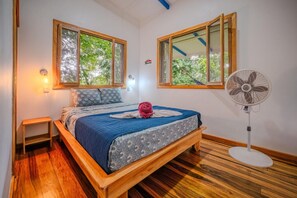 Une chambre à coucher avec un lit en bois, deux fenêtres, un ventilateur sur pied et une table de chevet.