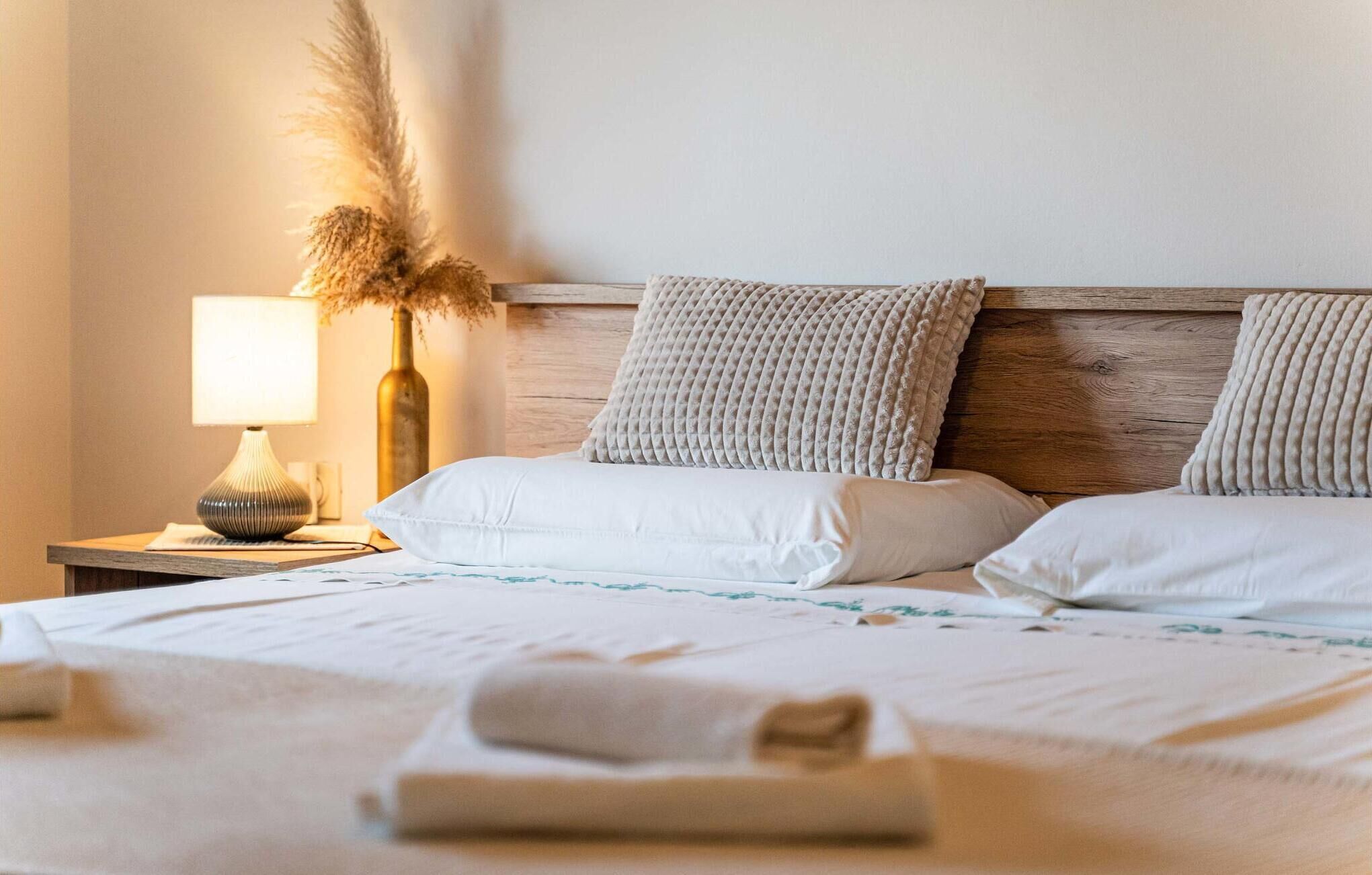 A neatly made bed with white linens and two pillows, a bedside lamp, and a vase with dried pampas grass.