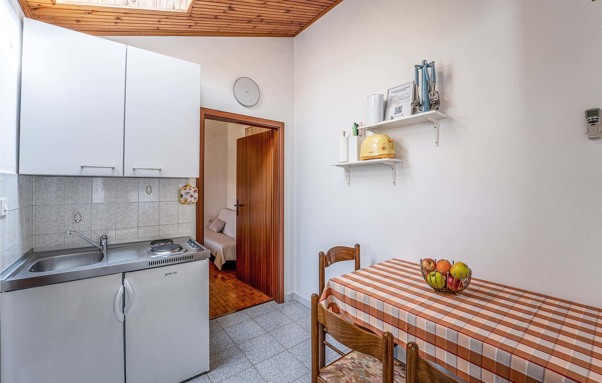 A compact kitchen with white cabinets, a stove, and a sink. There is a dining area with a table and chairs, and a small shelf with kitchen items.